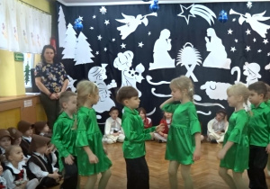 Na tle bożonarodzeniowej dekoracji tańczą dziewczynki w zielonych sukienkach i chłopcy w zielonych koszulach i czarnych spodniach.